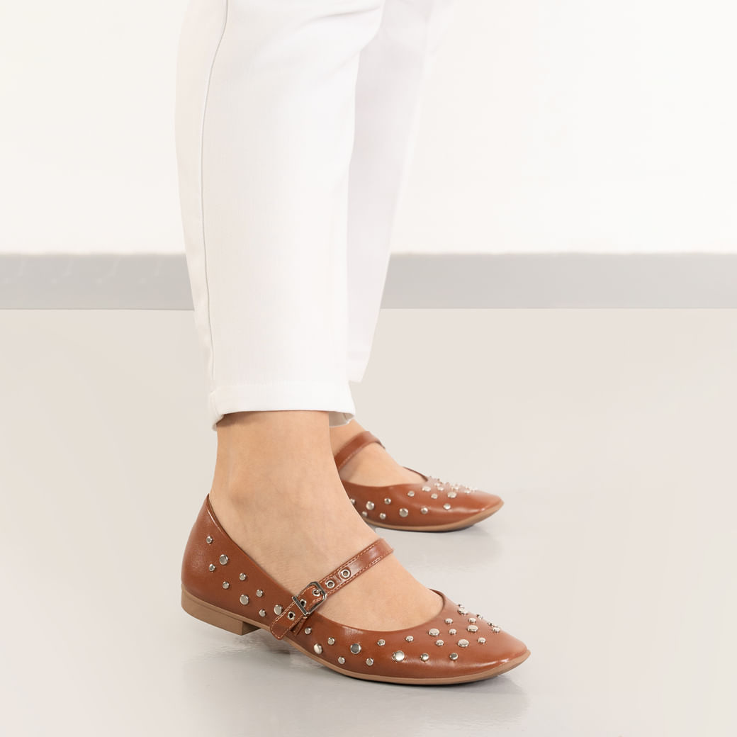 Sapatilha caramelo com bico arredondado, tira boneca com fivela metálica e tachas prateadas no cabedal, vista frontal com pé vestindo