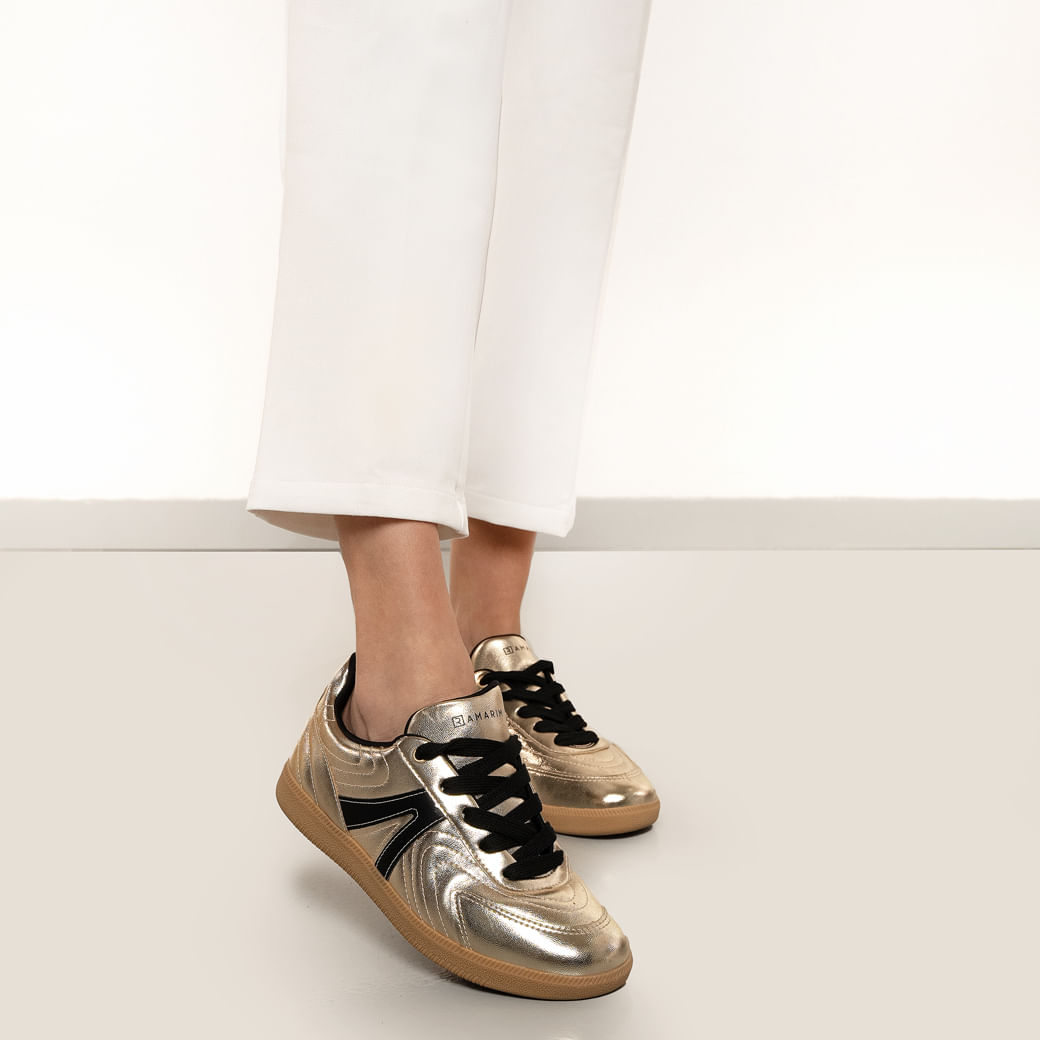 Close das pernas com tênis dourado metalizado matelassê e cadarço preto em fundo branco