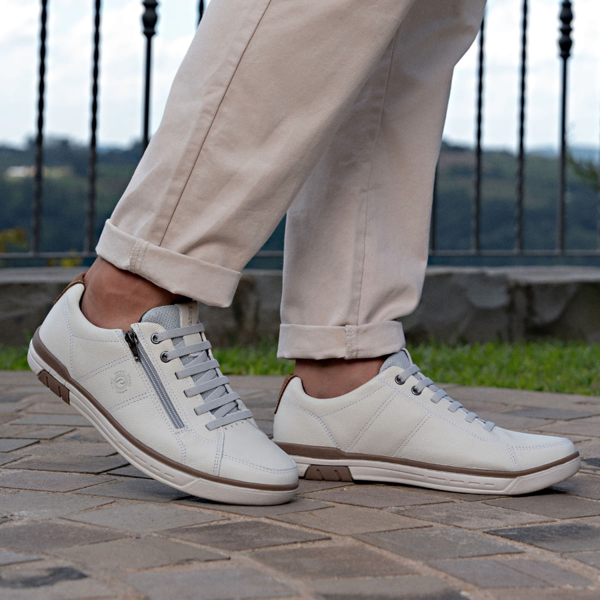 Tênis casual masculino branco com zíper lateral e detalhes marrons, em outro ângulo ao ar livre